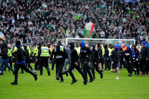 PA Media A number of football fans running across a football pitch wearing black clothing and face coverings.