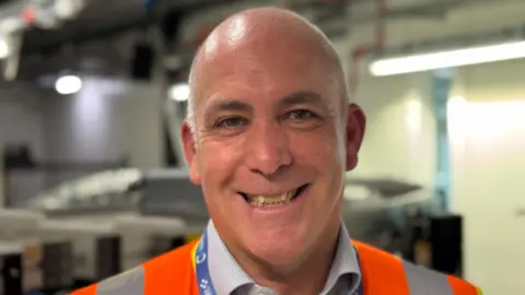 Stevie smiles to the camera, he's standing in front of a baggage carousel and is wearing a high vis jacket
