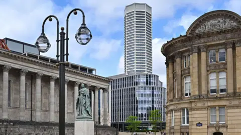 The Octagon Birmingham The Octagon towering over Birmingham's Chamberlain Square. It is gleaming white and has eight sides. There is a crown on top. It is a clear day with few clouds in the sky. We can see Birmingham's old-fashioned architecture, including its council house, clashing with the modern design of The Octagon.