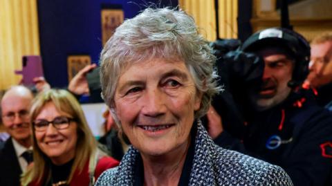 Catherine Connolly wears a woven blazer. She has short cropped grey hair and is smiling.