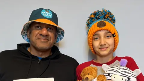 Jay Jayamohan Jay is wearing a black zip-up top and a blue hat with the Harry's Hat logo, while Anna is cuddling two cuddly toys and is wearing a grey and red shirt. She's also wearing an orange and blue Harry's Hat bobble hat