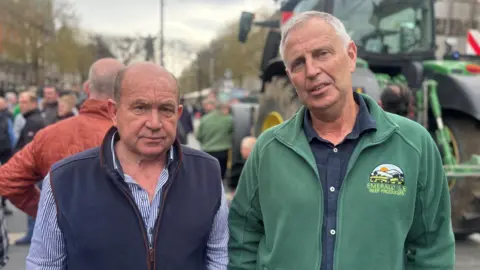 Two men are standing side by side looking into the camera. John Dallon is on the left. He has balding dark grey hair and is dressed in a stripe shirt with a navy fleece gilet on top. Eamon Curley has white hair and is dressed in a navy shirt with a green fleece on top. It has a stitched emblem on the chest with the words 'EMERALD ISLE BEEF PRODUCTS' below. The background is out of focus, but there is a large group of people gathered for the protest beside a green tractor. 