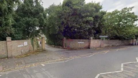 Road with entrance to brick wall lined driveway with dense trees either side.