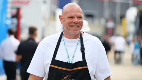 Tom Kerridge in a white t shirt and wearing a black chef's apron. There are people walking in the background. He is standing in a Formula One paddock.