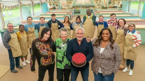 Love Productions/Channel 4/PA Paul Hollywood is standing holding a cake with raspberries on it and is stood with Noel Fielding, Alison Hammond and Dame Prue Leith. They are stood in front of a row of 12 contestants who are wearing brown aprons.