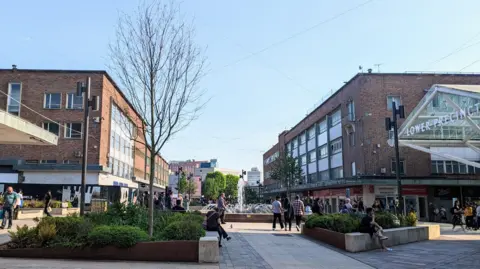 BBC Coventry city centre, with flower beds and a fountain surrounded by red brick shops. The flowerbeds have a mix of green bushes and trees while people sit on their edges and others wander through