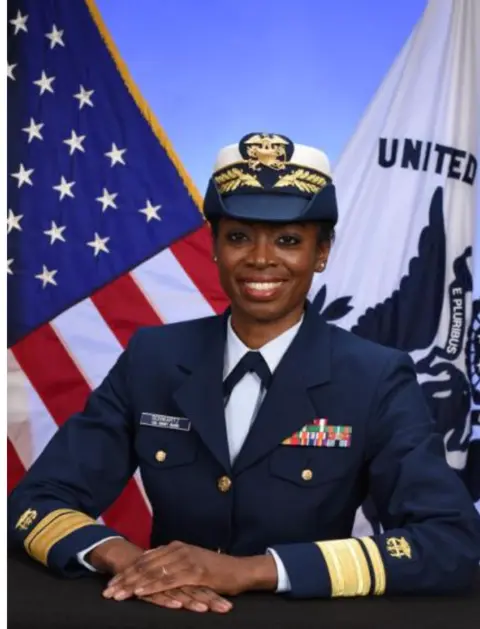 An official portrait of a woman in uniform with flags behind her. She is sitting and her hands are folded