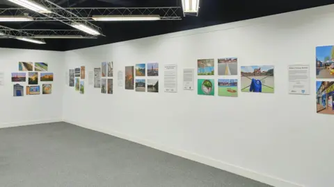 Martin Phillips/Historic England A wide shot of several clusters of photographs alongside caption boards, none of which are clearly visible but include a range of landscapes and close-ups of heritage features. They are displayed on a white wall with a lattice frame holding lighting above.