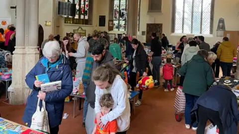 Sustainable Devizes Lots of people milling about inside a church where there are lots of tables with books, toys and homeware. In the foreground a family with a young boy are looking at some children's books, and an older lady is holding a pile of books.