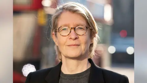 LGB Alliance Kate Barker looking direct to camera. She is standing outside on what appears to be a sunny evening. She is wearing a grey top under a black blazer-type jacket. She has blonde shoulder length hair, swept back over her ears and is wearing round-rimmed glasses with mottled amber and brown frames. A double decker red bus can be seen out of focus in the background with its headlights on.