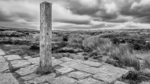 David Elder A black and white image of a carved wooden pillar on a rustic stone paved area, surrounded by rugged grassland in the Cambrian mountains on a cloudy day.