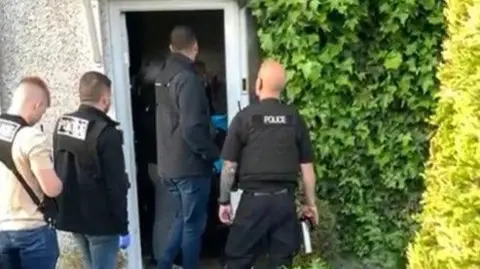 Four police officers can be seen entering the open door of a property, which has ivy growing down one side.
