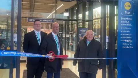 The three Reform councillors all wearing black jackets, white shirts and pale blue ties. Two are bald, while the third has short dark hair. The one in the middle is holding a pair of giant red scissors as they stand behind a Lidl branded ribbon which has the words 'new store' on it, with the glass doors of Lidl in Cwmbran behind them.