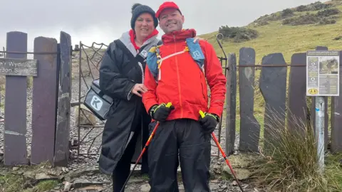 Lucy Chatting A woman wearing a black coat, bobble hat and with a bag, smiles as she stands next to a man with a red coat, hat and walking poles. They are standing in front of a gate on a footpath.