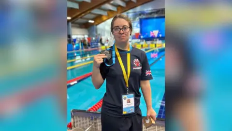 Family Handout A woman is standing in the middle of the frame, wearing a yellow lanyard around her neck and holding a medal. She is standing in front of a swimming pool.
