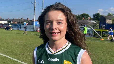 Paula McGivern has long wavy brown hair and is standing on a grass pitch. She is wearing a dark green short sleeved jersey with white and yellow trim. The top says o'neills in white and GAA on one side. A yellow club crest is on the other side.