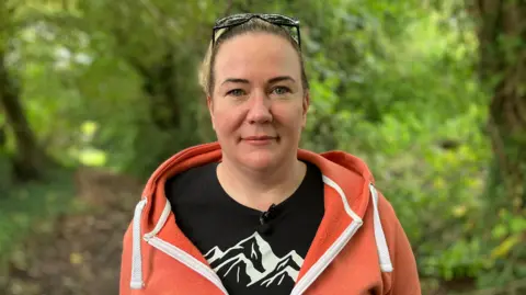 Helen is looking into the camera and is wearing sunglasses on her head. She has her hair pulled back and is wearing an orange hoody and a black t-shirt with a logo depicting a mountain. She's in a forest, but the leafy background is blurred.