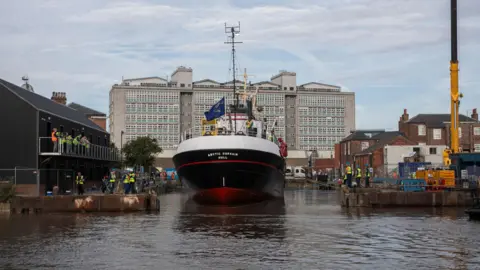 The trawler Arctic Corsair - a black-hulled boat with white upper works - enters a dock from the waters of the River Hull. People watch from the banks and the balcony of a black two-storey building. The skyline is dominated by a large concrete and glass building.