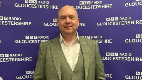 Cameron Thomas MP wearing a white shirt, green suit and waistcoat. He is standing in front of a purple wall with a repeating purple pattern of the BBC Radio Gloucestershire logo. 