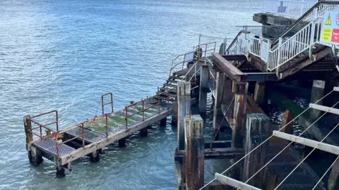 LDRS A platform connected the the pier via a staircase. The platform, a narrow jetty, is in visible disrepair. A metallic banister that extends around its perimeter is rusted and parts of it are missing. 