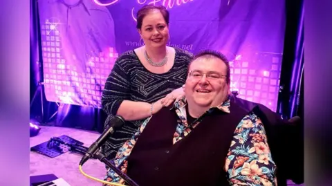 Stroke Association Stephen sits, while his wife Pauline stands and rests her hands on his shoulder. Stephen has brown hair which is slightly spikey, glasses and wears a bright hawaaian shirt, with a black waistcoat, while Pauline, who has short red hair, wears a sparkly top, silver necklace and gold hoop earrings.
