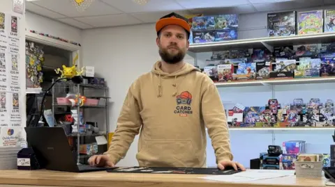 Card Catcher Shop Sam Jackway, owner of Card Catcher Shop in Bristol, stands at the counter, with Pokemon cards and other collectibles on shelves behind him