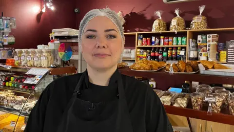 A woman stands in front of a bakery counter packed with treats in various shapes, colours and sizes. She is wearing a white hair net and a black apron over a black top.