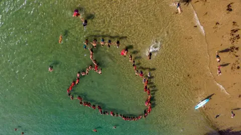 People stood in seawater near the short of a beach. They are wearing red and and are stood in the shape of a heart. The beach is sandy. The water is clear and blue. 