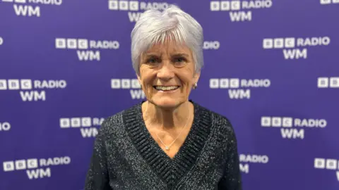 A woman with short white hair and wearing a necklace with a black jumper, stands and smiles in front of a purple background with the white BBC Radio WM logo all over it.
