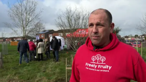 Matt Knight stands to the right facing the camera in front of a group of people who are gathered around a new tree in an open green space. He is wearing a red hoodie with the charity's logo in white on the front. He has short brown hair