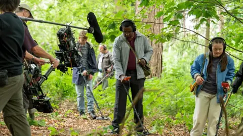 BBC/House Productions/Sam Taylor A group of six people stand in woodland, one of them is filming with a large camera, and a boom mic is also visible