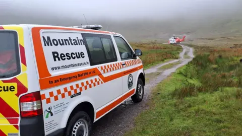 C Sexton SLMRT response vehicle with ICG Rescue 118 helicopter on standby near the base of the mountain. The vehicle is white with orange pattern. The helicopter is white and orange. There is a path and grass in the foreground with mountains in the background. It is cloudy.