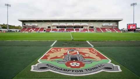 Getty Images A general view of Sixfields Stadium, home of Northampton Town. A stand with red seats can be seen on the opposite side of the pitch. Directly in front of the camera, on the ground, is the club badge emblazoned on the turf. 
