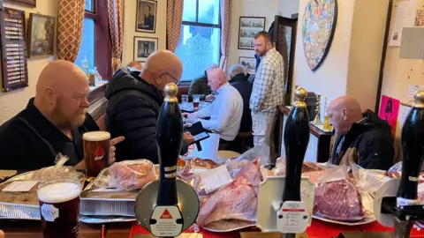 General view of a pub and there is meat laid out on the bar. Behind it are several people gathered around, chatting and drinking.