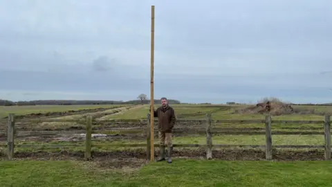 A man in a brown coat and light brown trousers, wearing wellies, is standing next to a tall post on farm land.