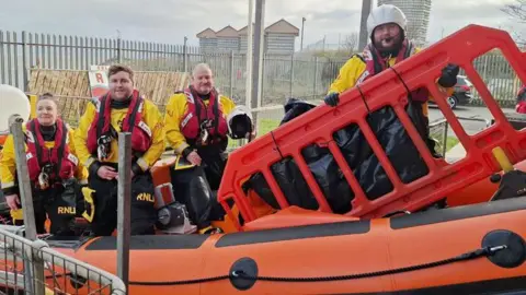 RNLI/Tom Collins Four crew members in white waterproof suits and red life jackets stand on an orange inflatable boat. One of them is holding a large red plastic fence