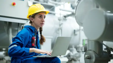 Getty Images Young female engineer wearing a yellow hard hat and blue overalls works on a laptop on an industrial site 