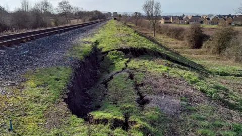 An area of grassland next to a railway line has collapsed. There are trees and a row of houses in the background.
