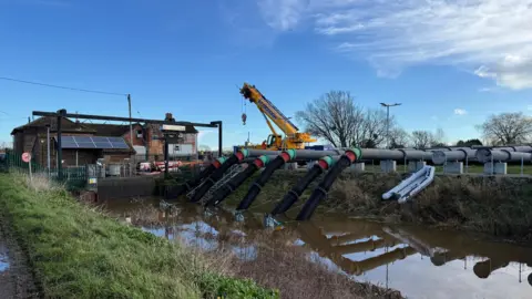 A number of large black pumps lead in to a body of water. A yellow crane is visible in the background.