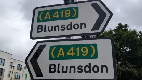 Anna Bird/BBC Two smaller official road signs - the upper one points to the right, saying it goes to the A419 and Blunsdon. The one just below points to the left and also says it goes to the A419 and Blunsdon. A grey sky above.