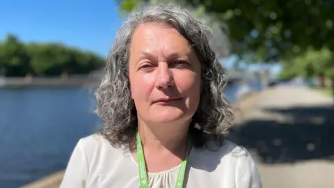 A profile shot of Fiona Needham, a Trading Standards, boss standing on the Victoria Embankment along the River Trent. She has short, curly grey hair, and is wearing a white T-shirt and a green lanyard