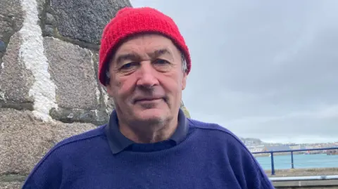Man in his 60s wearing a blue Guernsey and a knitted woollen red hat