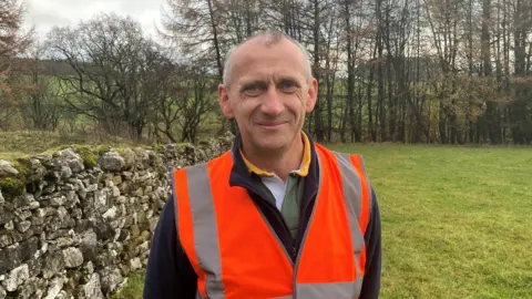 Ian Hutchinson is standing beside a dry stone wall in a field. He is smiling towards the camera wearing an orange hi-vis vest over a black fleece. In the background trees without autumn leaves can be seen against a background of the Cumbrian fells. 