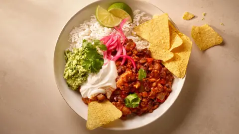 A white bowl containing rice, tofu chilli, guacamole, lime and tortilla chips.