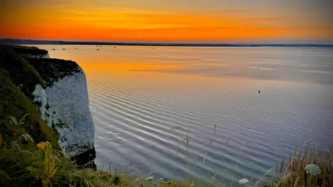 White cliffs to the left of the image sees ripples across a flat sea spreading up to the horizon where the sky is bright orange graduating up to yellow as the sun rises