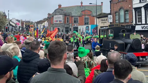 A bustling crowd, donned in festive green attire and hats, lines a city street to watch a vibrant St. Patrick's Day parade. Central to the scene is a float resembling a black steam locomotive, labeled 'St. Patrick's Express,' promoting the Downpatrick & County Down Railway. The parade route is flanked by various buildings, including shops like 'VOGUE' and 'O2.' 