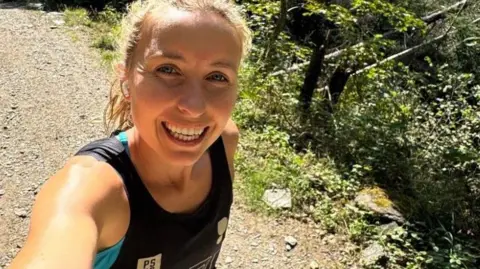 Beth Rainbow Beth Rainbow in a selfie style photo. She has blonde hair and is wearing a running singlet while on a dirt track.