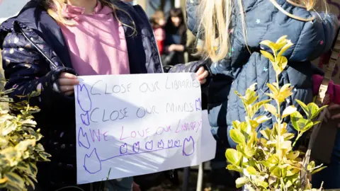 A girl is holding a sign, which reads 'Close Our Libraries, Close Our Minds, We Love Our Library'