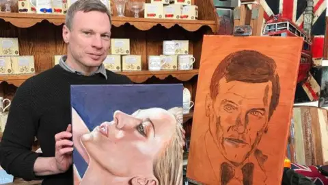 Tristan Copeland A man with dark grey hair wearing a black sweater. He is holding a painting of a woman's head. She has blonde hair and is tipping her head back. Next to him, on an easel, is another piece of art which is entirely orange in colour and has a black charcoal drawing on it of a man