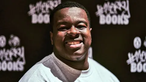 Will Fahy Abaka Debrah, winner of the #Merky Books prize, smiling and wearing a grey top. Behind him is branding which reads "Merky Books" in white letters.